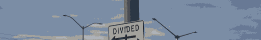 A sign on a lightpole reading "divided" with an arrow going to the left. THe sign has been divided in half. There are tops of streetlights in the background.